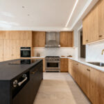 Wide-angle photograph of a contemporary luxury kitchen layout featuring an oversized island, light oak cabinetry, and wide aisles designed for efficient movement in a multi-cook household.
