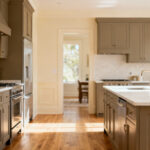A professionally photographed modern kitchen interior with warm greige cabinets, light wood flooring, and soft white walls, demonstrating a perfectly harmonized color palette through consistent undertones, creating a serene and cohesive atmosphere. Bright natural light fills the room, accentuating the subtle interplay of colors.