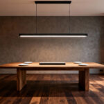 A sleek black linear suspension light fixture hangs centered over a long rectangular wood dining table in a modern dining room, demonstrating uniform and shadow-free task lighting.