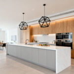 A wide view of a minimalist open-concept modern kitchen featuring a large white quartz island, light gray handleless cabinets, and light oak wood storage, designed to accommodate multiple users and collaboration.