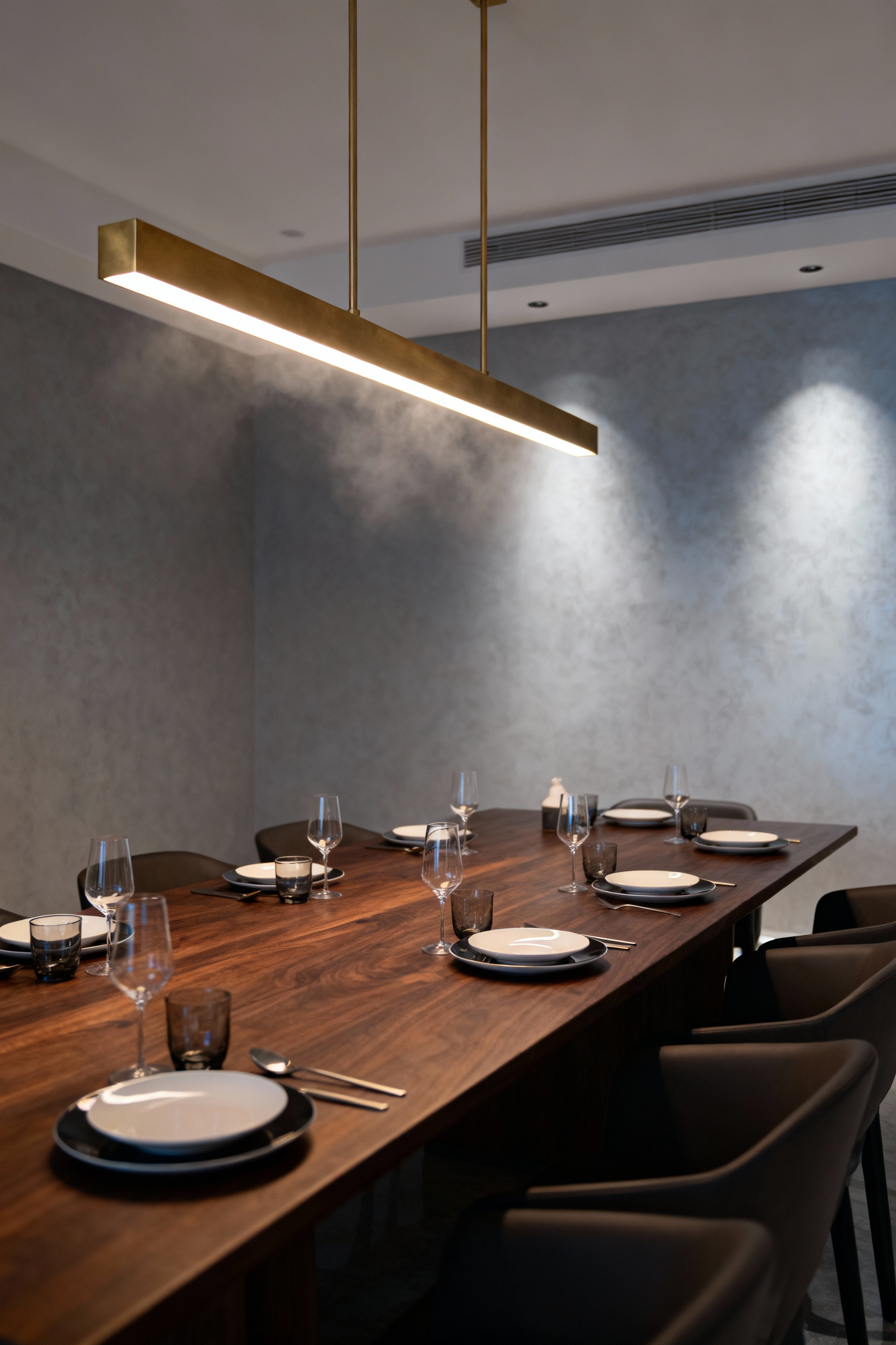 A luxurious dining room featuring a long dark wood table illuminated by a modern brass linear LED chandelier, showcasing the rich, warm ambiance achieved through smart voice-controlled lighting.