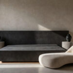 A modern, high-design bedroom interior where minimalist furniture is treated as sculptural art, featuring a monumental platform bed, asymmetrical concrete nightstands, and dramatic, high-contrast lighting emphasizing form.