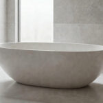 A photograph of a sleek, modern, freestanding bathtub made of volcanic limestone composite material set in a minimalist luxury bathroom with bright natural light.